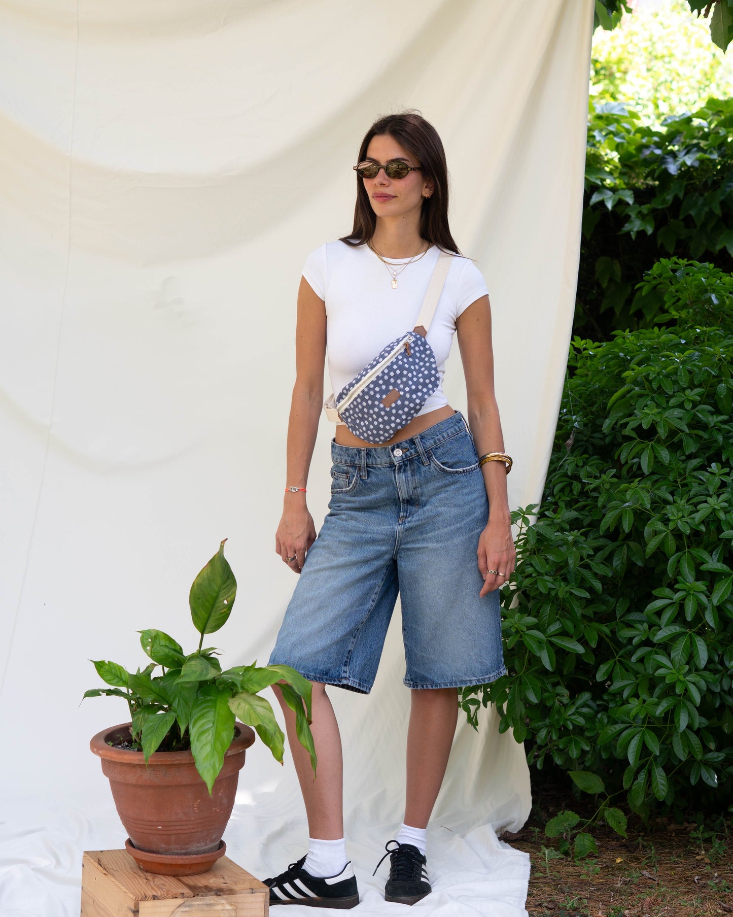 Sac banane écoresponsable et upcyclé sur une jeune fille debout avec un fond blanc et des fleurs sur le coté. sac banane couleurs bleu jeans avec des fleurs marguerites et une sangle blanche parfaite pour l'été,