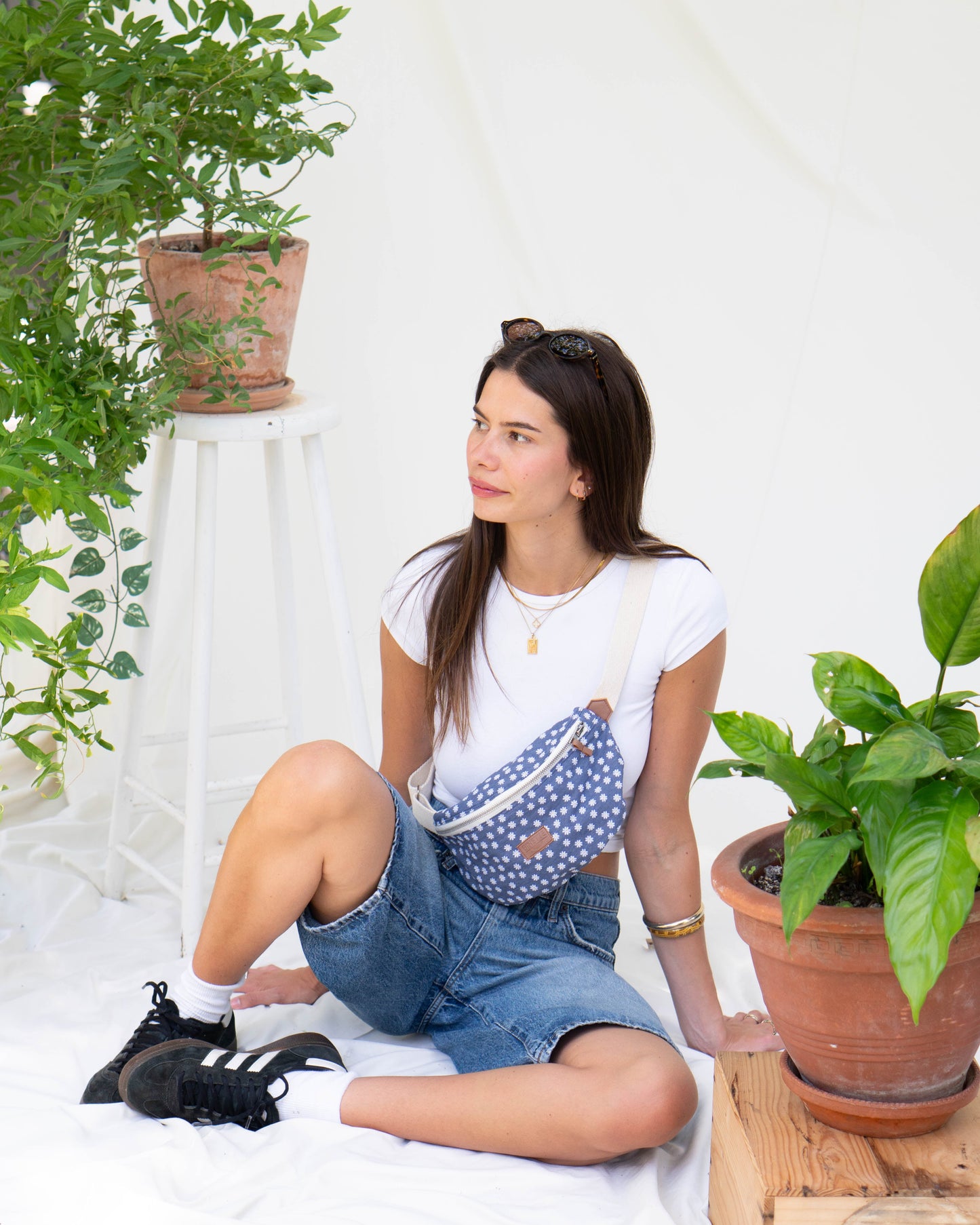 Jeune femme brune avec un t-shirt blanc et un short jean assise sur un fond blanc avec des fleurs portant un sac banane bleu jeans fonce avec des fleurs marguerites dessinés dessus ecoresponsable et recyclé