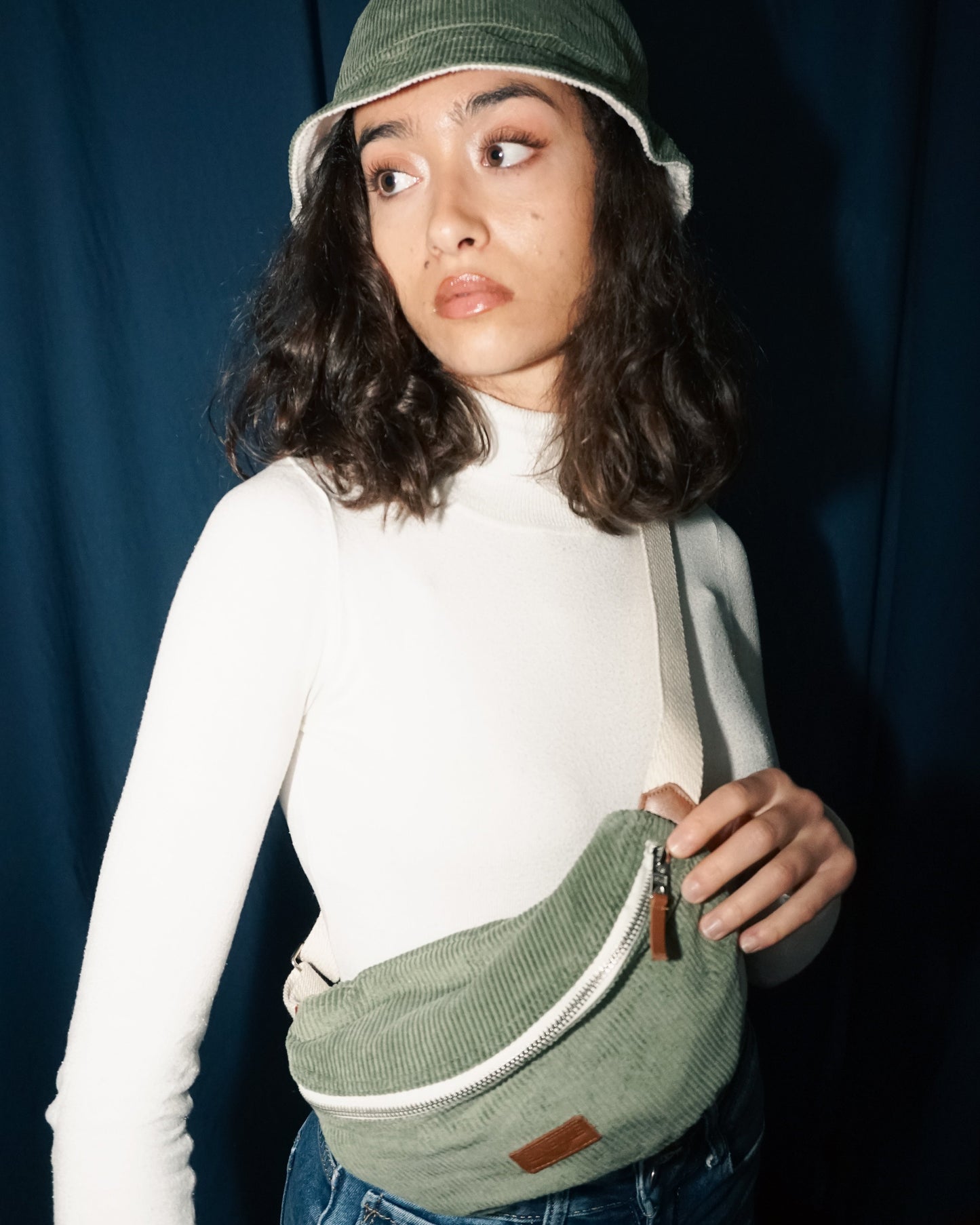 Femme portant un bob vert velours côtelés avec un sac banane assorti et écoresponsable et upcyclé avec un fond bleu marine et un pull blanc