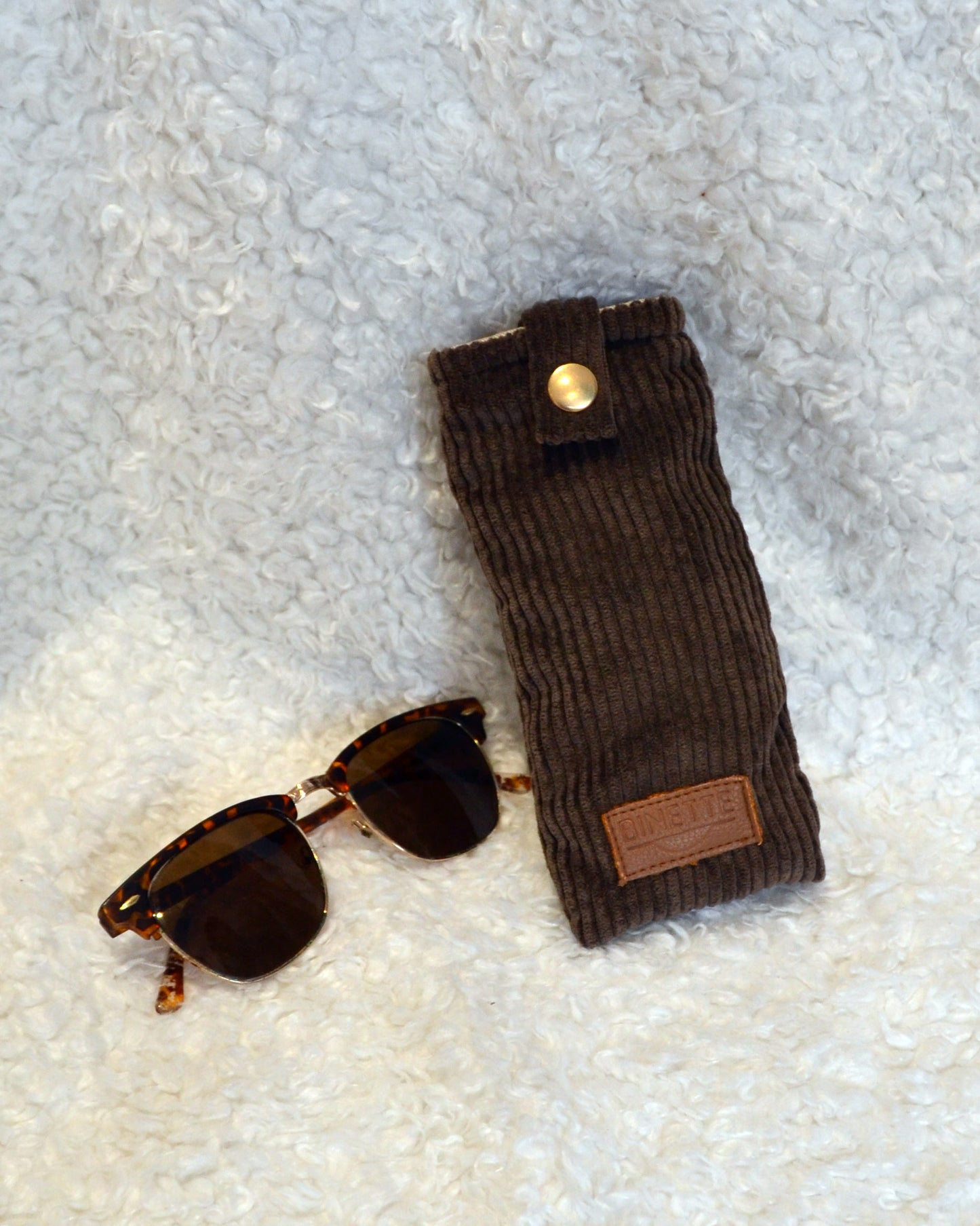 Étui à lunette marron chocolat velours côtelé avec paire de lunette marron sur moumoute blanche.