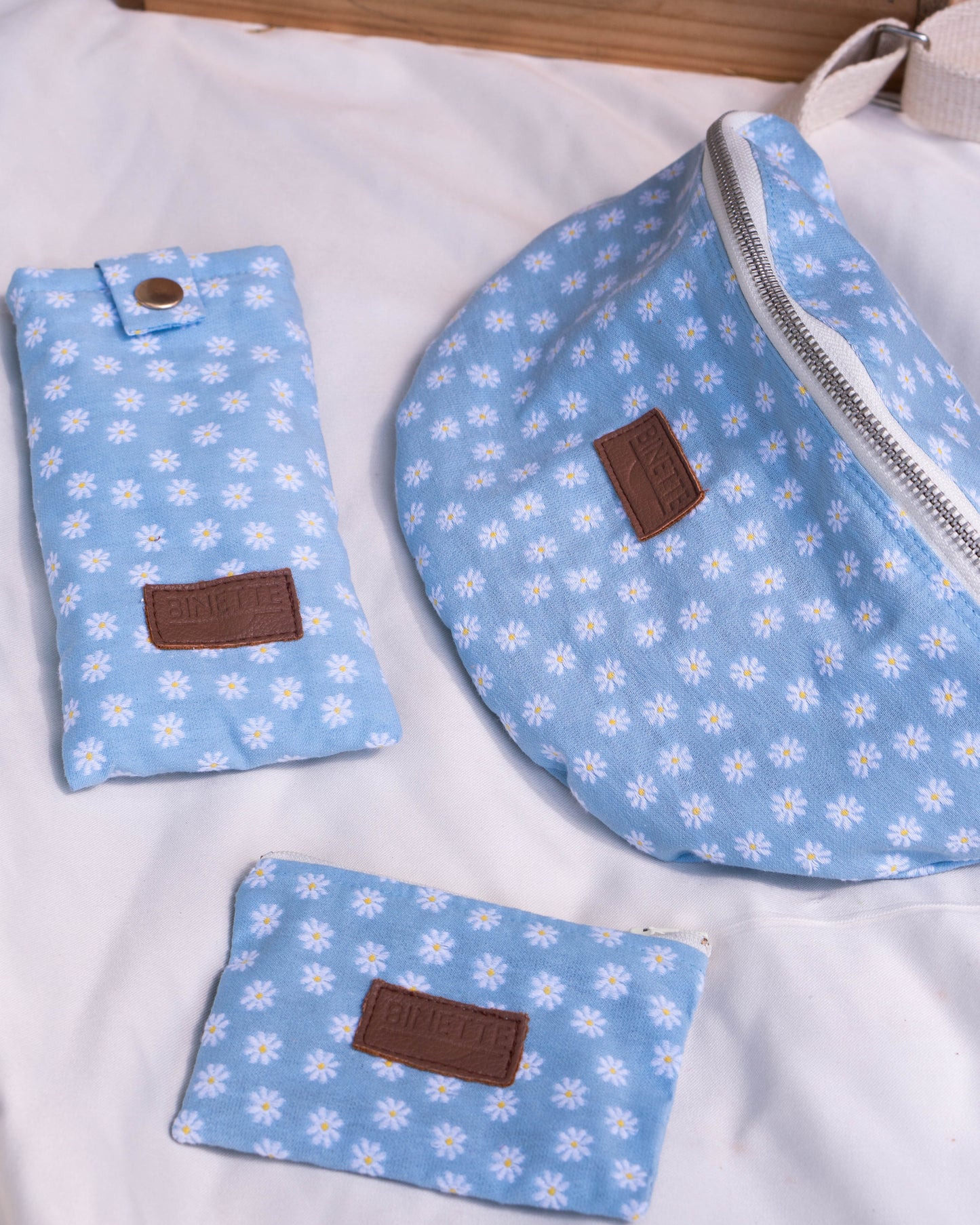 Packshot d'un sac banane écoresponsable et recyclé sur un fond blanc avec pochette à fleurs et étui à lunettes à fleurs couleur bleu ciel et motif marguerite. Pack parfait pour idée cadeau femme et jeune maman