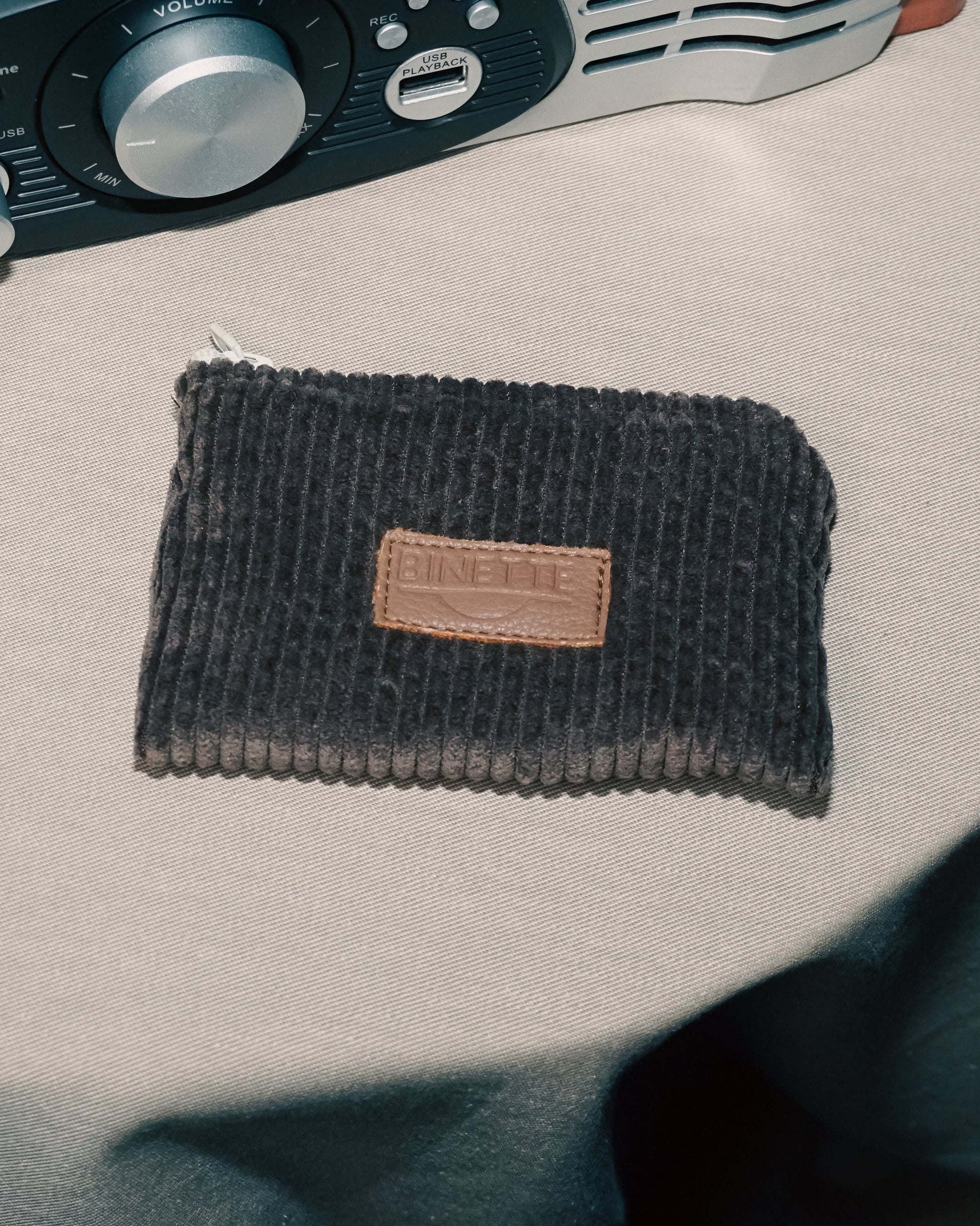 Porte-monnaie marron chocolat en velours côtelé avec logo Binette en cuir et fermeture blanche posé sur une table a côté d'une enceinte.