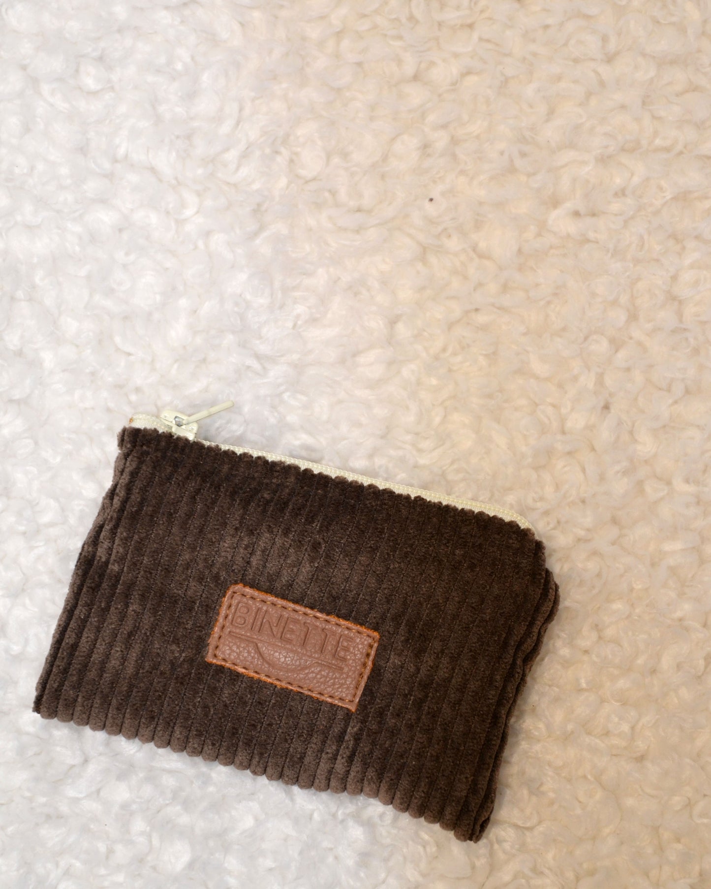 Porte-monnaie marron chocolat en velours côtelé avec logo Binette en cuir et fermeture blanche posé sur une table à côté d'une enceinte.