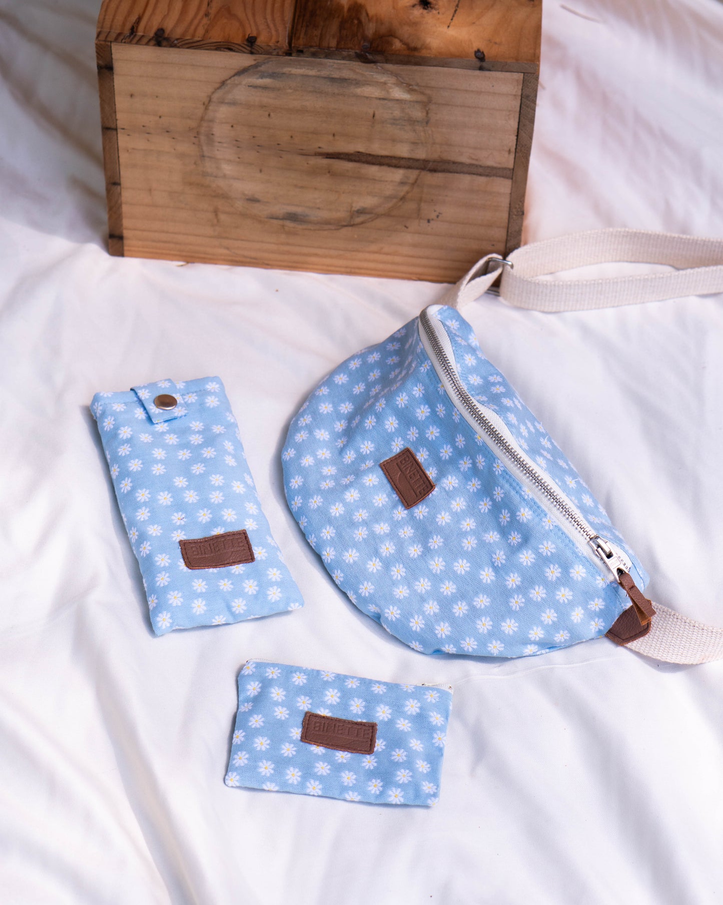 Packshot d'un sac banane écoresponsable et recyclé sur un fond blanc avec pochette à fleurs et étui à lunettes à fleurs couleur bleu ciel et motif marguerite. Pack parfait pour idée cadeau femme et jeune maman