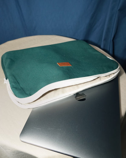 Pochette d'ordinateur en velours côtelé vert sapin posée sur une table aux côtés d’un laptop. Alliant élégance et protection, elle est conçue à partir de matériaux upcyclés et fabriquée en France pour une touche à la fois stylée et responsable.