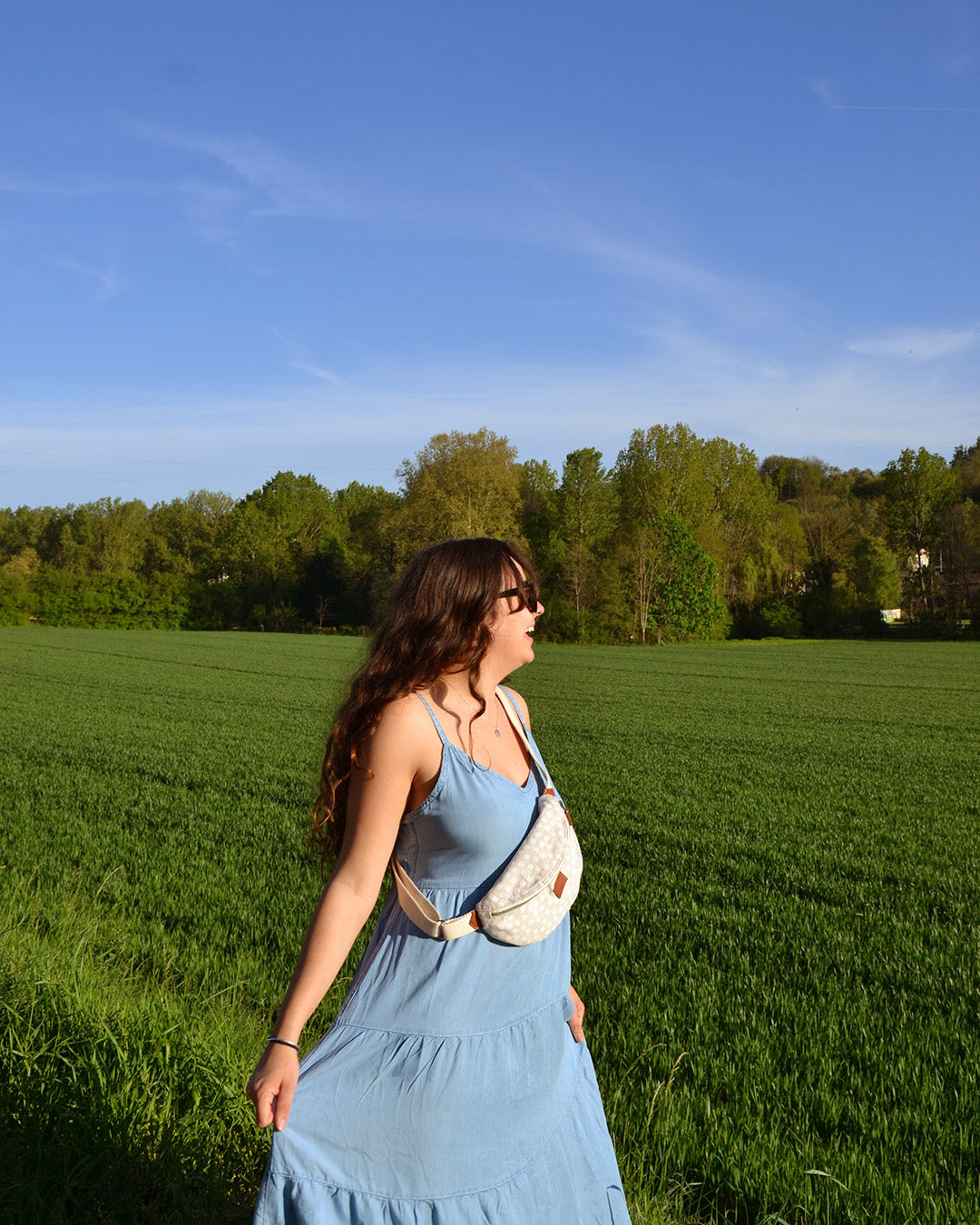 Cette image captivante montre une femme portant la Binette Marguerite Beige à la taille, ajoutant une touche de décontraction bohème à son look estival. Son sourire radieux témoigne du confort et de la praticité de ce sac, qui lui permet de garder l'essentiel à portée de main tout en restant élégante. La Binette Marguerite Beige devient ainsi un accessoire incontournable pour les journées bien remplies, offrant à celle qui la porte un style décontracté et tendance.