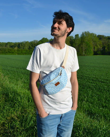 Dans cette image, un homme porte avec style un sac banane bleu orné de multiples marguerites blanches. L'accessoire ajoute une touche de fraîcheur et de couleur à sa tenue décontractée. Pratique et tendance, ce sac est parfait pour une journée en ville ou une sortie entre amis. On y retourve sur le devant le logo de Binette.