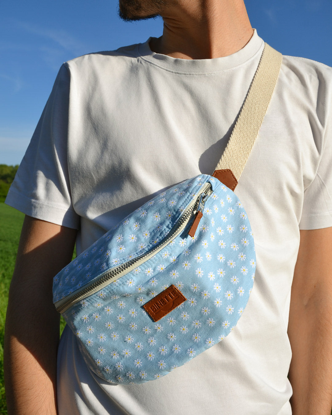 Dans cette image, un homme porte avec style un sac banane bleu orné de multiples marguerites blanches. L'accessoire ajoute une touche de fraîcheur et de couleur à sa tenue décontractée. Pratique et tendance, ce sac banane est parfait pour une journée en ville ou une sortie entre amis.