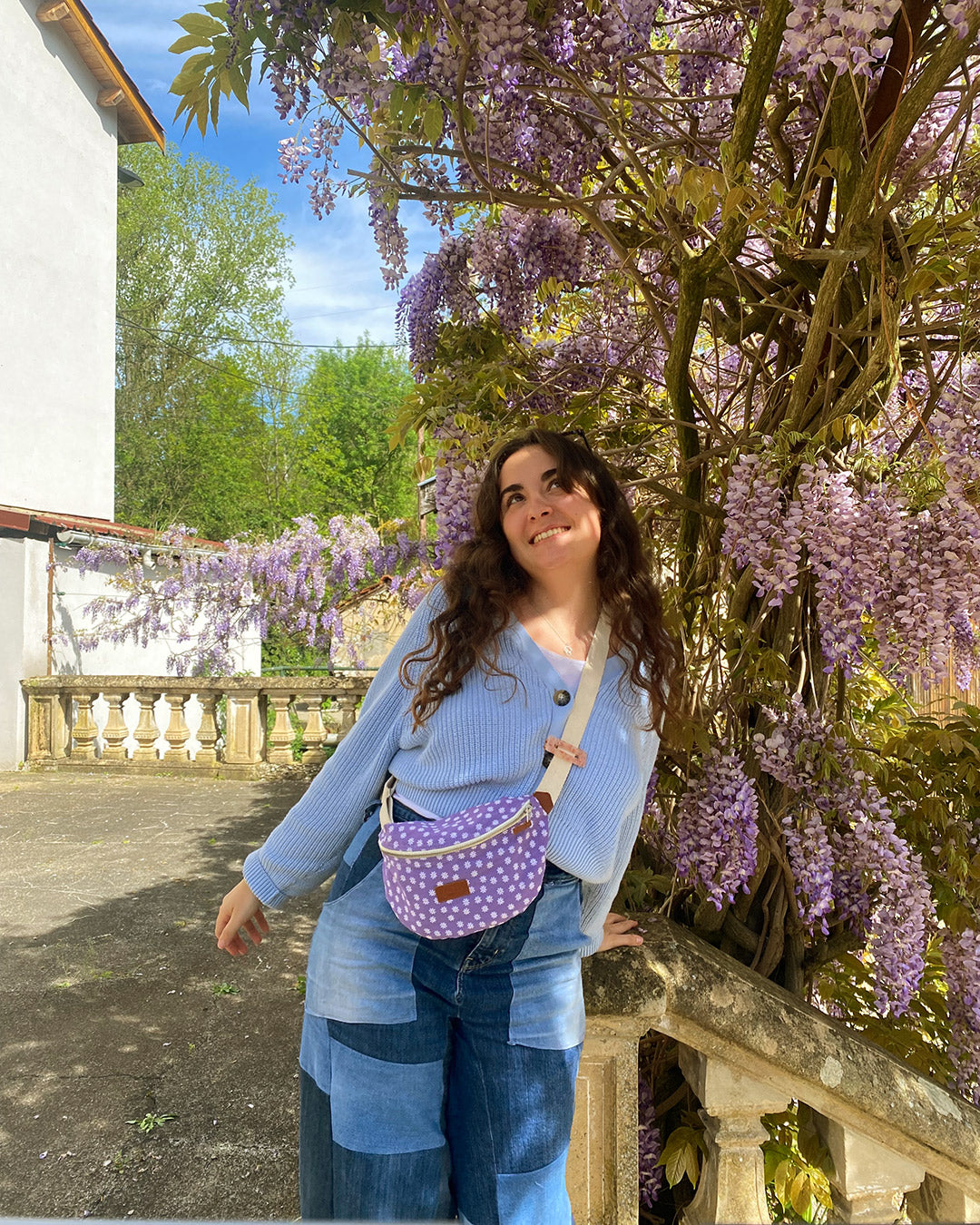 Dans cette image, une femme souriante porte la Binette Marguerite Violette en bandoulière pendant une balade en ville. Ses mains libres témoignent de la praticité de ce sac, qui lui permet de garder l'essentiel à portée de main tout en restant élégante.