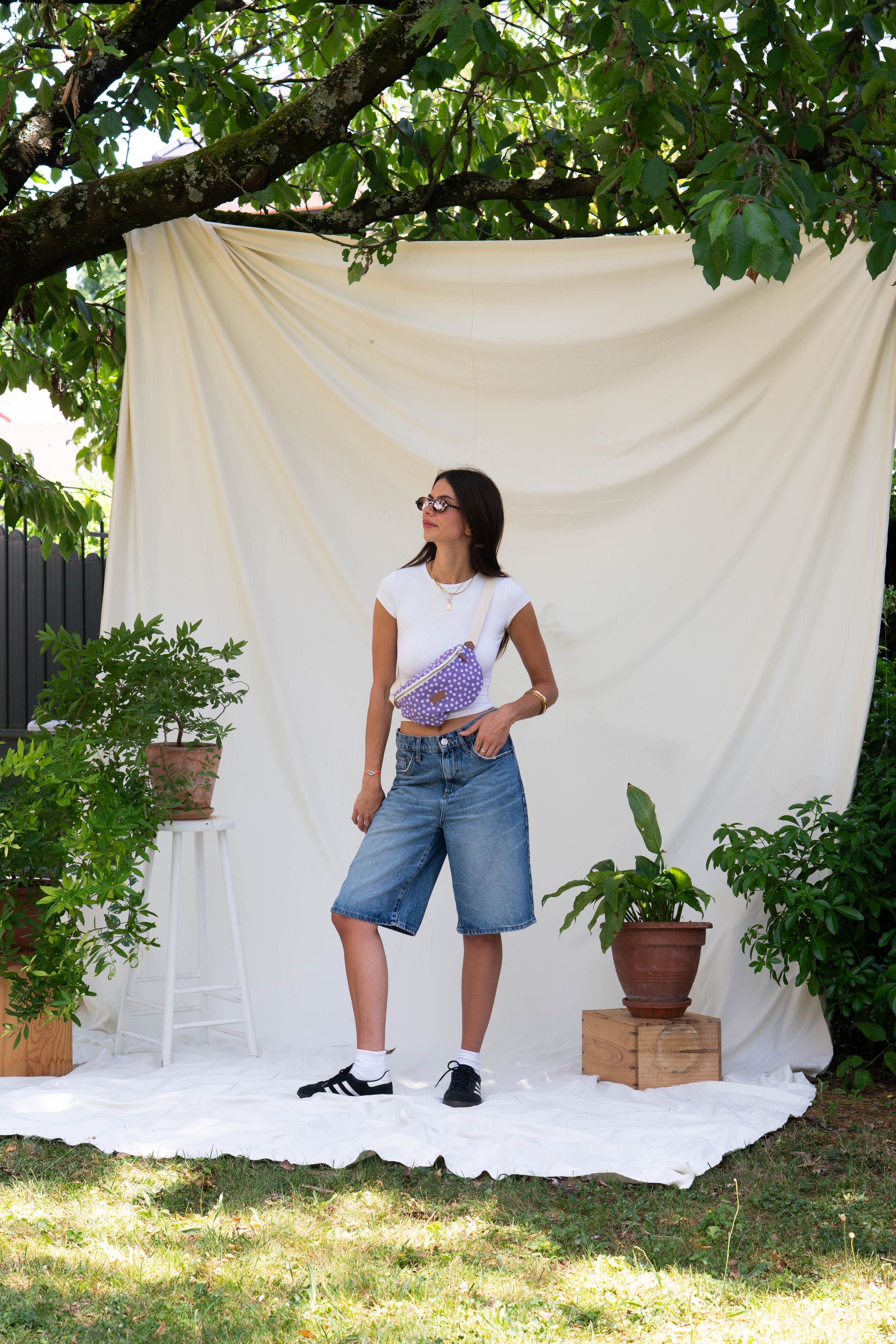 Dans cette image, la modèle debout porte la Binette Marguerite Violette en bandoulière en taille L. Banane élégante, bohème, chic et parfaite pour idée cadeau coloré. 